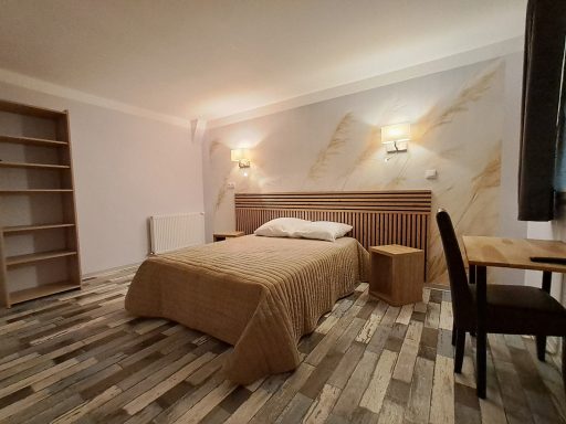 Chambre simple avec lit, table, chaise et étagères en bois. Lumières douces au mur.